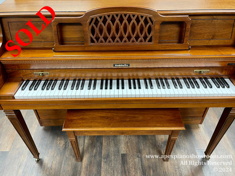 A classic Baldwin upright piano with a polished brown wood finish and intricate lattice music stand visible above the keyboard, set against a dark wood flooring background.