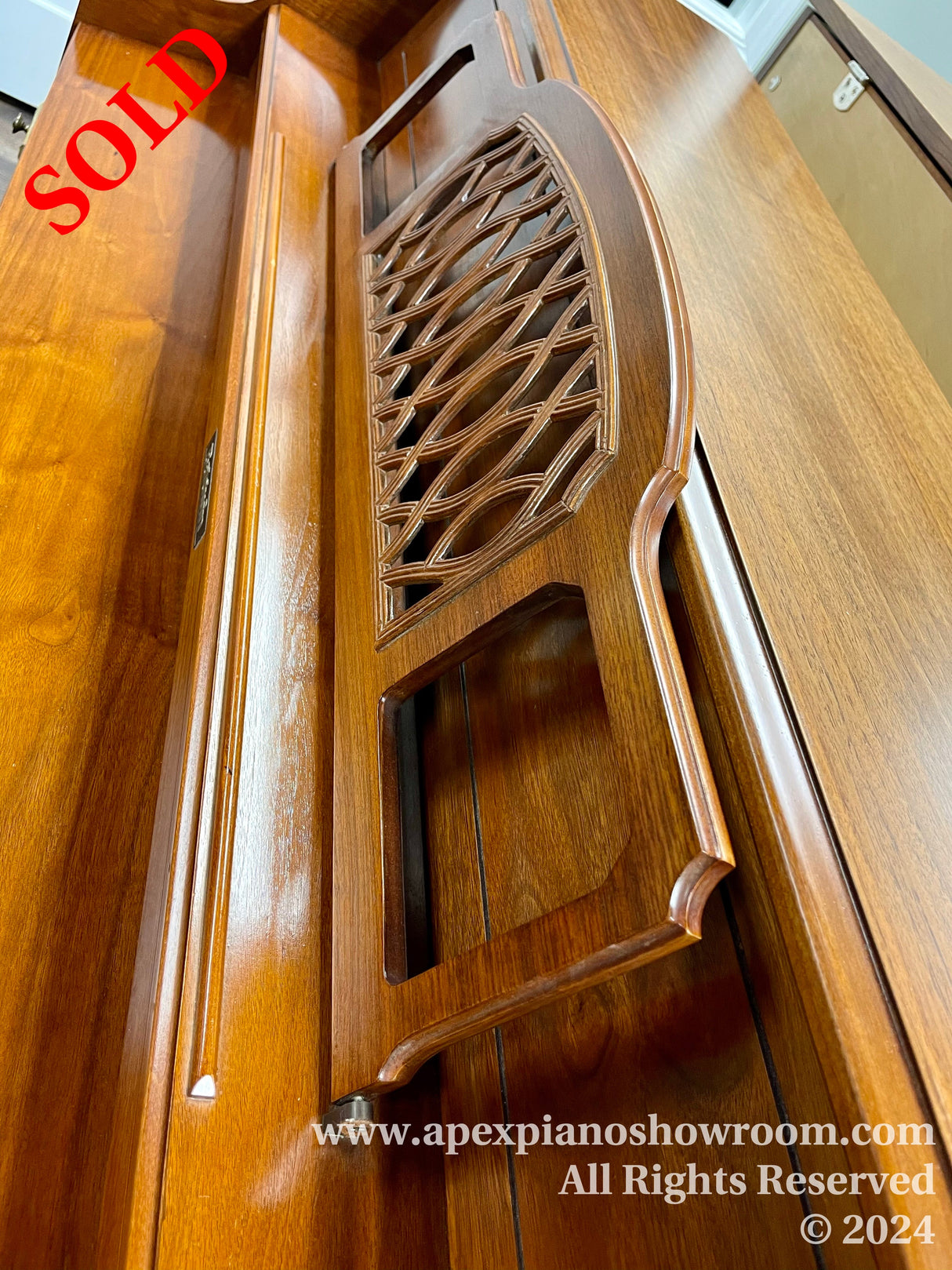 Close-up view of a wooden piano music desk with intricate lattice design, piano fallboard with handle, and the edge of the piano lid showing polished wood finish.