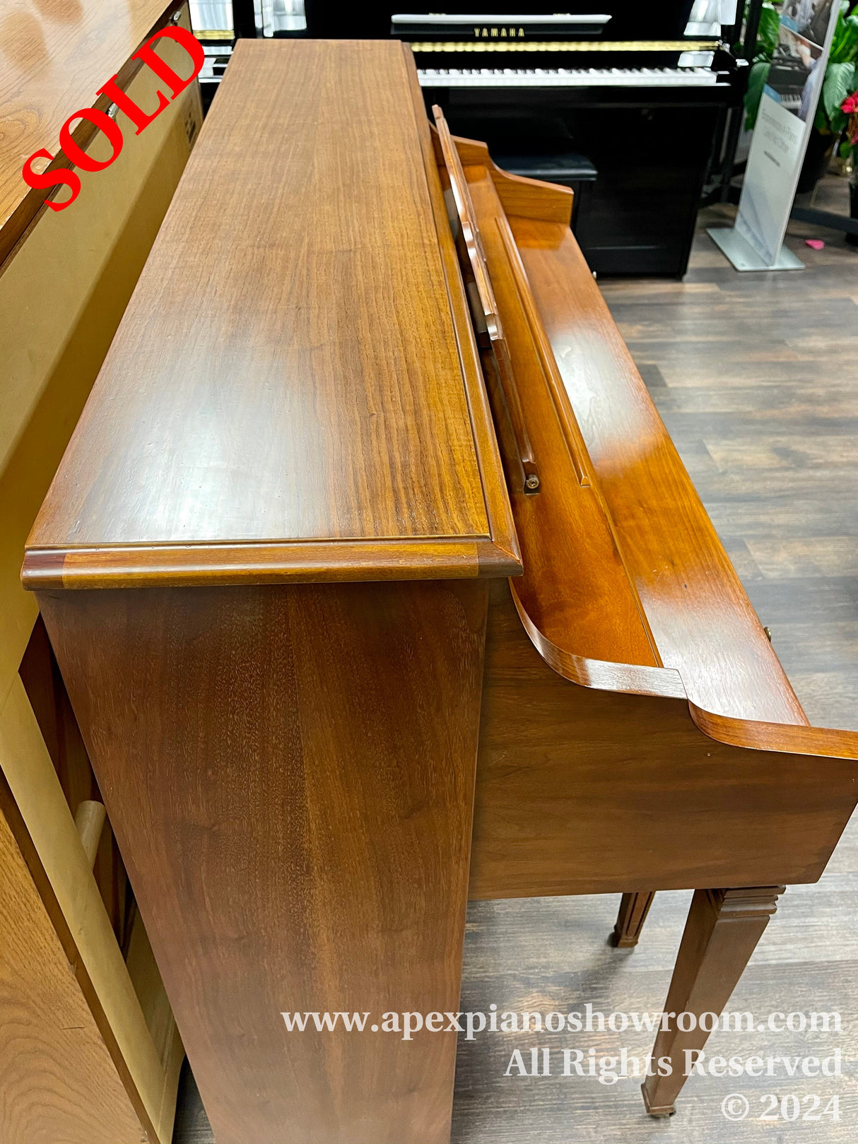 A close-up view of two upright pianos with rich wooden finishes displayed in a showroom — the closest piano has a smooth, reflective brown surface and is positioned in front of another piano with a black and white keyboard visible in the background.