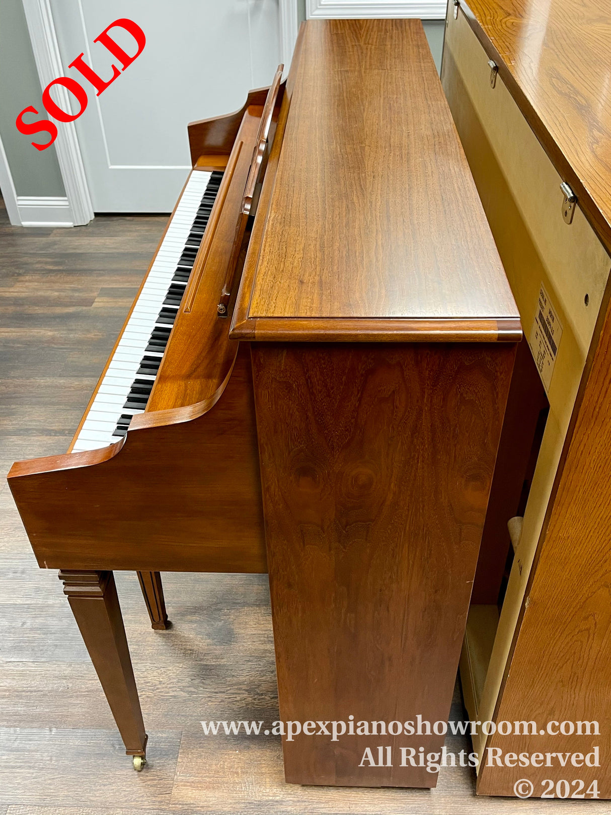 An upright piano with a brown wooden finish on display in a showroom, featuring a hinged top cover revealing ivory and black keys, with piano legs and pedals visible, set against a wood-patterned floor and light grey walls.