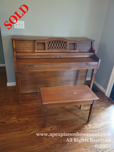 A Baldwin spinet piano with matching bench in a home setting, featuring a walnut finish and traditional decorative music desk, displayed on a hardwood floor.
