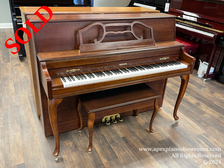 A classic Baldwin upright piano with a wooden finish, featuring a closed top with decorative music rest and elegant Queen Anne style legs, situated in a showroom with multiple pianos visible in the background.