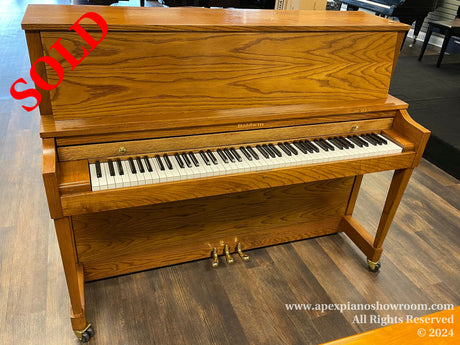 A Baldwin upright piano with a wood finish is displayed on a showroom floor, featuring a closed keyboard lid and brass caster wheels for easy movement.