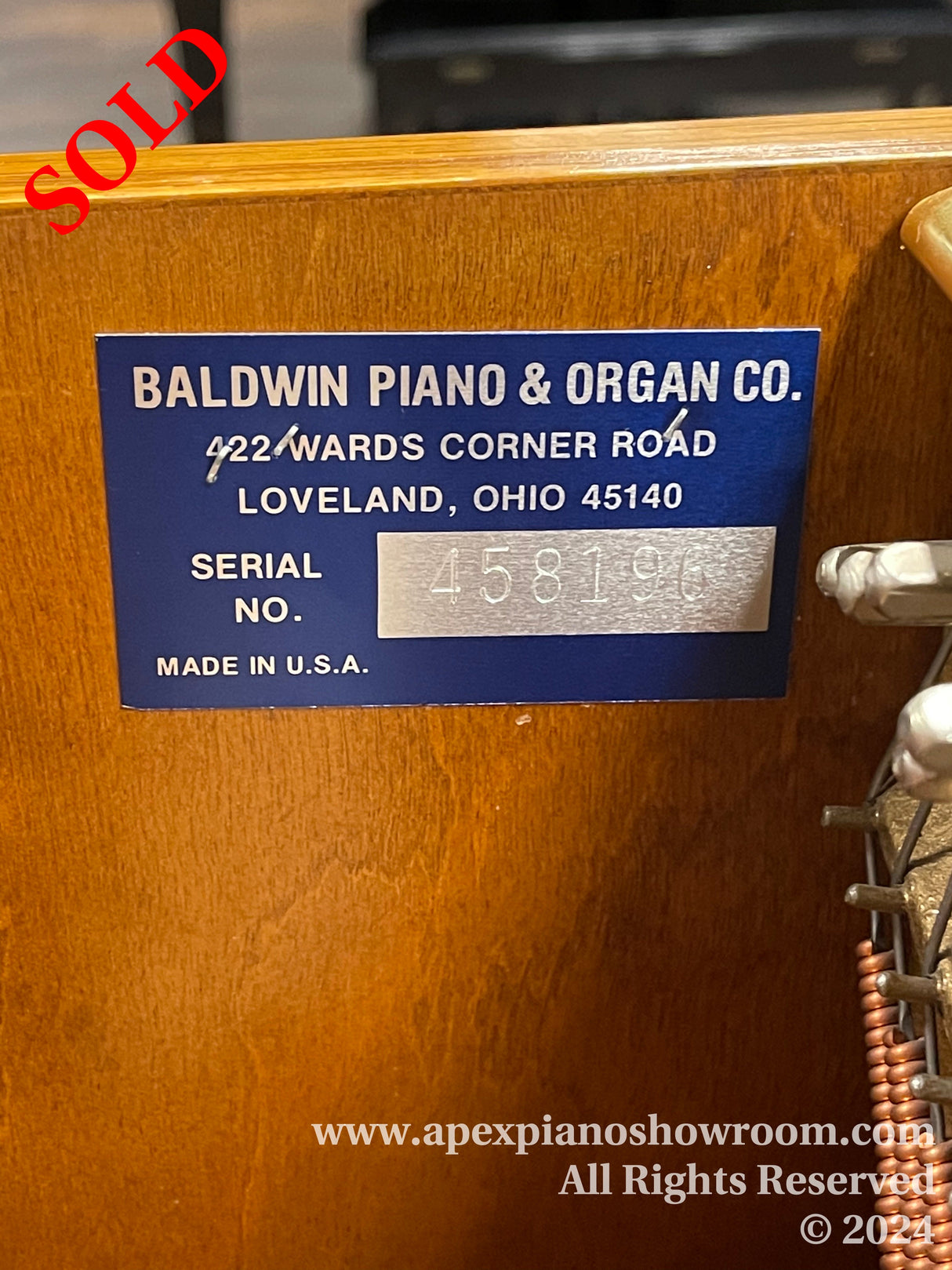 Manufacturers label on a Baldwin piano showing the company name, address in Loveland, Ohio, and serial number 458196, indicating the pianos identification details.