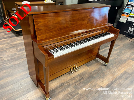 A polished mahogany Baldwin upright piano with a closed top, featuring brass hardware and situated in a piano showroom with wood-patterned flooring.