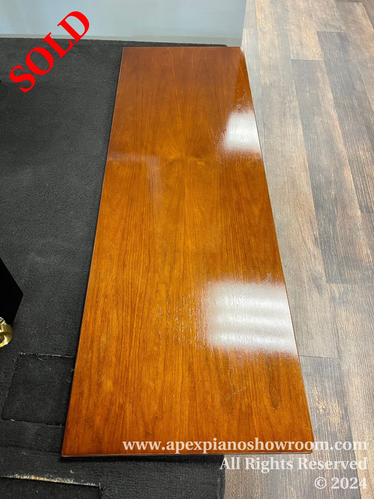 A glossy finished piano fallboard with rich mahogany color, resting on the floor against a black mat and grey wood-patterned flooring.