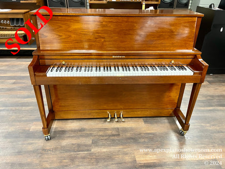 A Baldwin upright piano with a polished wood finish and brass caster wheels set in a showroom with wood laminate flooring.