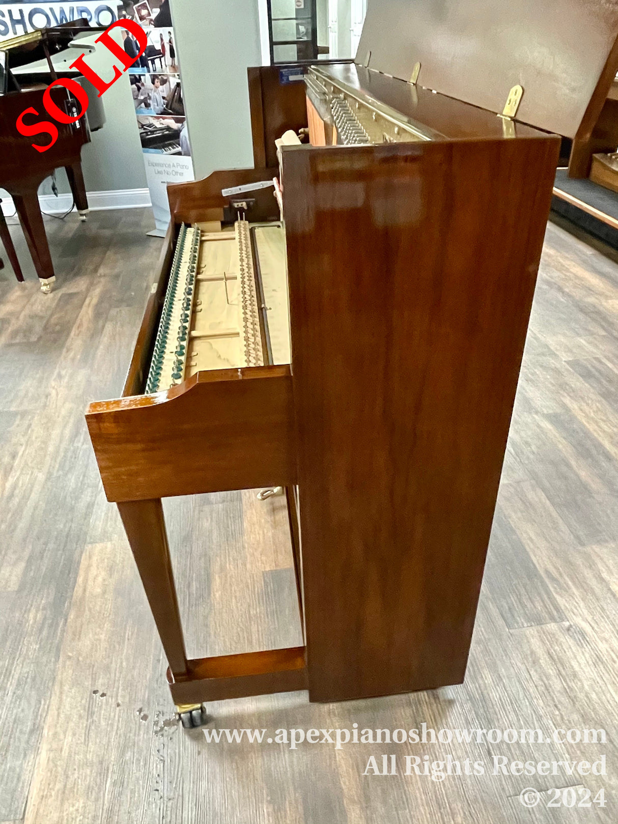 An open upright piano with its mechanism exposed, positioned in a piano showroom with hardwood flooring — a polished mahogany finish is prominent on its surface.