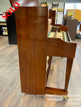 A wooden upright piano situated in a showroom, with its fallboard open revealing the keys, set against a floor with wood flooring.