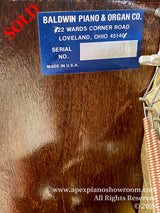 A close-up view of a Baldwin pianos serial number plate, which is affixed to a wooden surface with text that includes Baldwin Piano & Organ Co., 422 Wards Corner Road, Loveland, Ohio 45140, with the serial number 457200 clearly displayed. The plate indicates that the piano is made in the U.S.A. The background shows the wooden texture of the piano and part of the instruments copper-colored tuning pins.