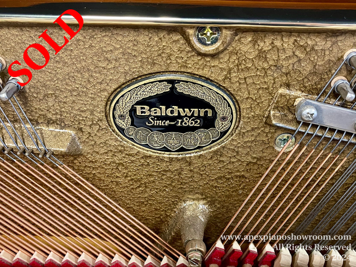 Close-up view of a Baldwin pianos interior showing the brand emblem, copper wound bass strings, and the gold-colored cast iron plate with a textured finish.