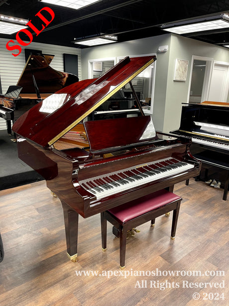 A glossy grand piano with mahogany finish and matching bench showcased in a piano showroom with other pianos in the background.