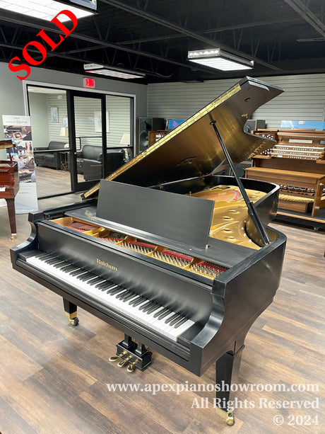 A Baldwin grand piano with a glossy black finish and gold interior is positioned open on the showroom floor, showcasing its keys and intricate internal strings and hammers, against a backdrop of other pianos and a seating area intended for customers or performers.
