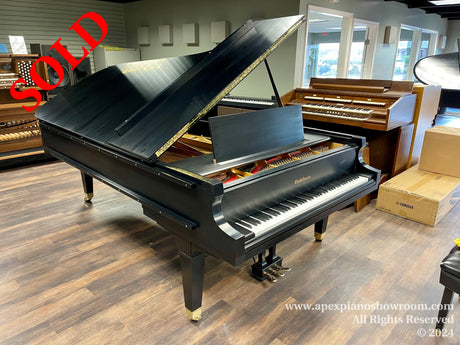 A glossy black grand piano with lid open on display in a piano showroom, surrounded by other pianos and musical instruments, with a wood flooring and soft lighting environment.