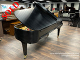 Black grand piano with open lid prominently displayed in a piano showroom, surrounded by various other pianos in a well-lit indoor setting.