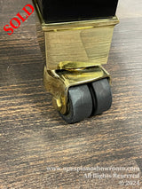 Close-up view of a piano caster wheel and leg, showing brass housing and black dual rubber wheels on a wood floor, typically used for easier movement and to protect the flooring from damage.