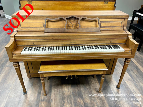 A wooden upright piano with an open top lid displaying intricate wood patterns and a music desk, set in a well-lit showroom with a matching bench.