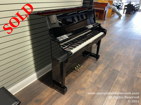 A glossy black upright piano positioned on a wooden floor in a well-lit showroom with various pianos in the background.