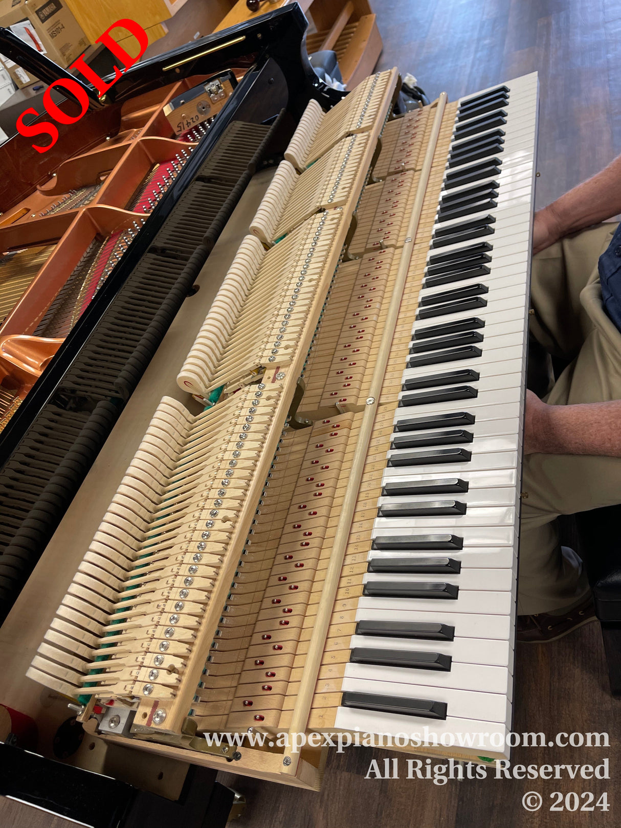 Grand piano with its action removed and placed next to the keyboard, showcasing the hammers and dampers, in a piano showroom with hardwood floors.