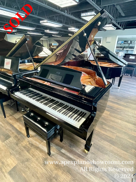 Black grand piano with an open lid, displaying its shiny black finish and interior strings, set in a wood-floored showroom with other pianos in the background.