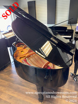 Glossy black grand piano with open lid showing strings and hammers, displayed in a showroom with other pianos in the background and wooden flooring.