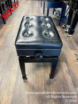 A black, tufted, adjustable piano bench with a storage compartment, positioned in a showroom with wooden flooring next to a polished black piano with caster cups under its legs.