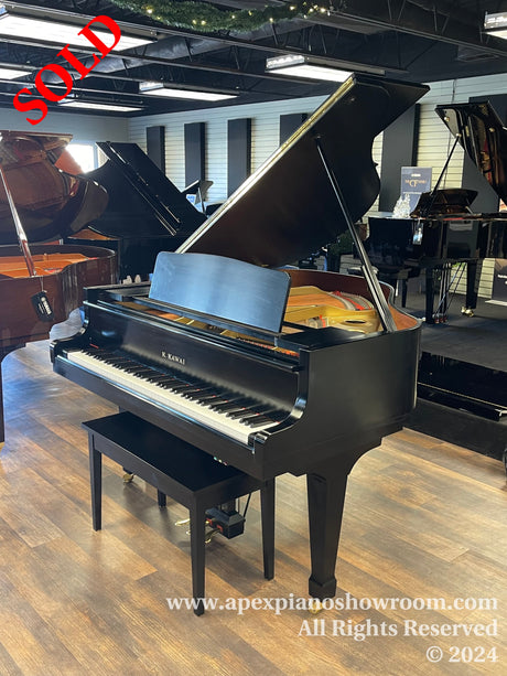 A shiny black Kawai grand piano on display in a showroom with its lid open, surrounded by other grand pianos, against an interior backdrop with a striped accent wall and recessed lighting.