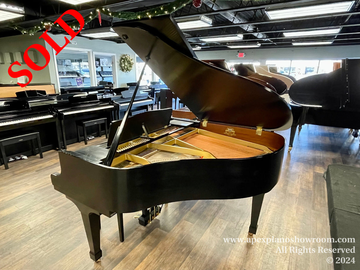 An open grand piano with a glossy black finish and exposed golden interior mechanics is prominently featured in a showroom surrounded by various closed black grand pianos on a wood-style floor.