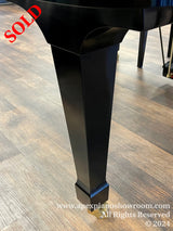 A close-up view of a shiny black piano leg with a sleek design, positioned on a wooden floor in a showroom setting, with a reflection of overhead lights visible on the polished surface.