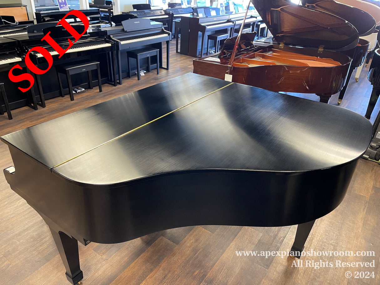 A shiny black grand piano with a closed lid is prominently displayed in a showroom with various other pianos in the background, set on a wooden floor, indicating a wide selection available for customers interested in high-quality pianos.