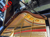 A grand piano with its lid open, revealing its intricate gold-colored interior mechanics, including strings and hammers, with a focus on the high-tension steel strings and copper wound bass strings, set in a showroom with other pianos in the background.