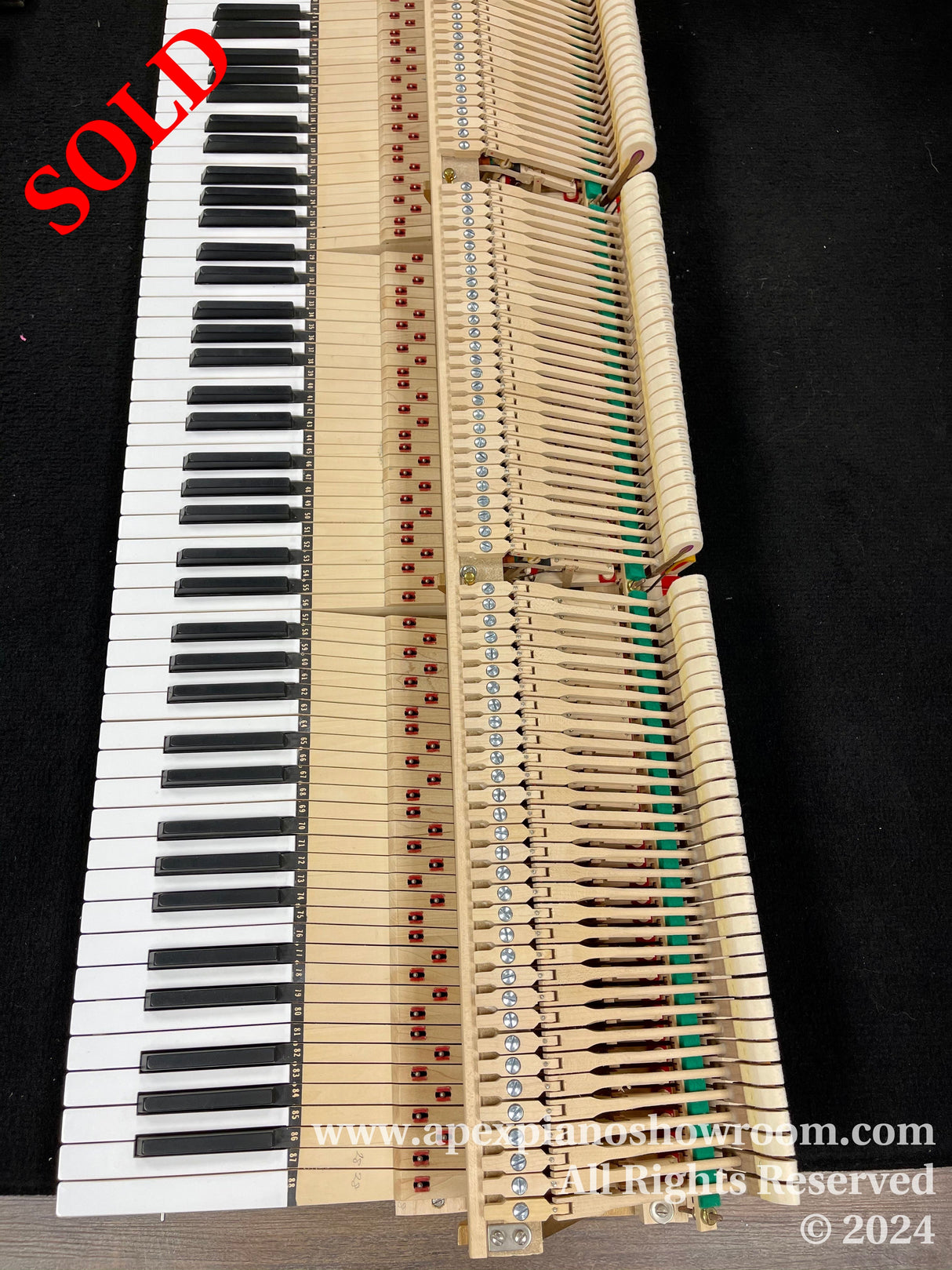 A close-up view of piano keys along with the internal hammer mechanism of a disassembled piano, showing the intricate array of hammers and dampers with felt, arrayed in a diagonal fashion across the image with a black background.