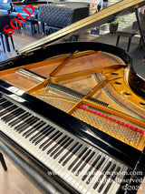 A grand piano with its lid open, showcasing its interior mechanics including strings and hammers, with a focus on the black and white keys of the keyboard. The piano is situated in a showroom with other pianos visible in the background.