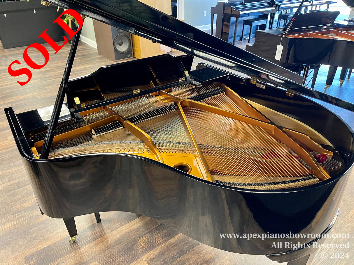A glossy black grand piano with its lid open, displaying intricate internal mechanisms including strings and hammers, situated in a well-lit showroom with wood flooring and other pianos in the background.