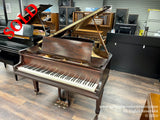 A brown grand piano with its lid open, showcasing the strings and hammers inside, situated in a showroom with a black grand piano in the background.