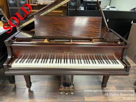 A professional brown wood-grain grand piano with the lid open, showcasing its internal strings and hammers, set against a background of a music showroom with audio equipment and a black office chair.