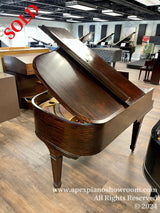 A glossy, dark wood grand piano with its lid open, showcasing the intricate internal strings and hammers, located in a well-lit piano showroom with multiple pianos on display in the background.