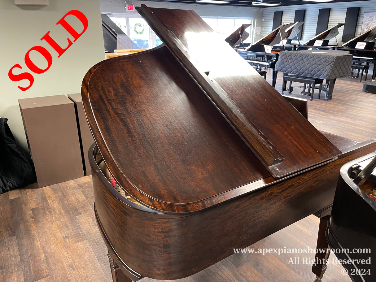 A grand piano with a rich, wood finish is prominently displayed in a showroom with various other pianos in the background, highlighting the variety and selection available for musicians and enthusiasts.