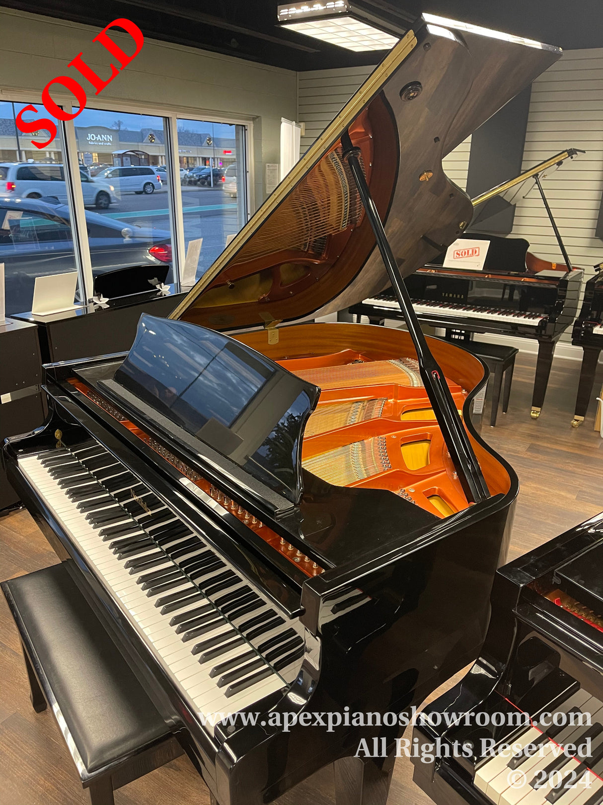 A high-gloss black grand piano with its lid open, showcasing the intricate internal string and hammer mechanism, placed inside a piano showroom with a bright interior and additional pianos in the background.
