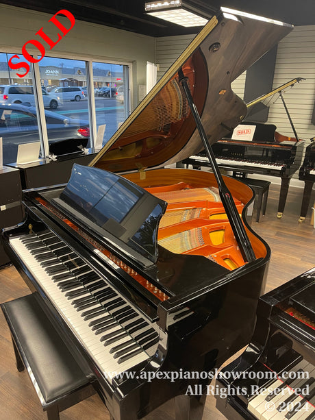 A high-gloss black grand piano with its lid open, showcasing the intricate internal string and hammer mechanism, placed inside a piano showroom with a bright interior and additional pianos in the background.