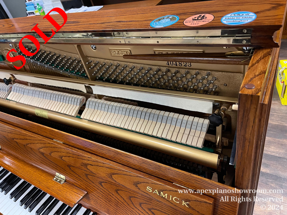 Interior view of a Samick upright piano showing the strings, hammers, and dampers, with a visible logo and serial number, set inside a woodgrain finished cabinet with the fallboard open.