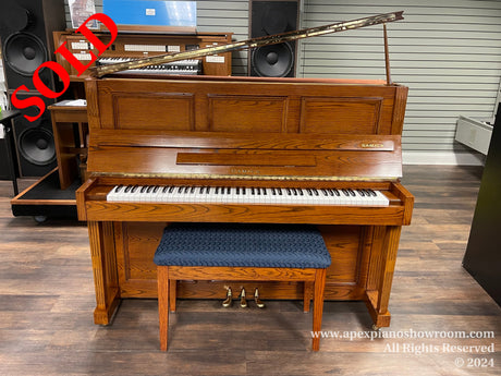 An upright Samick piano with a wooden finish, matching bench, and lid propped open, displayed in a showroom with speakers and other audio equipment in the background.