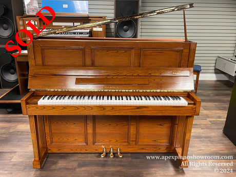 A Samick upright piano with a polished wood finish and an open lid, showcasing the pianos interior strings and hammers, situated in a showroom with musical equipment in the background.