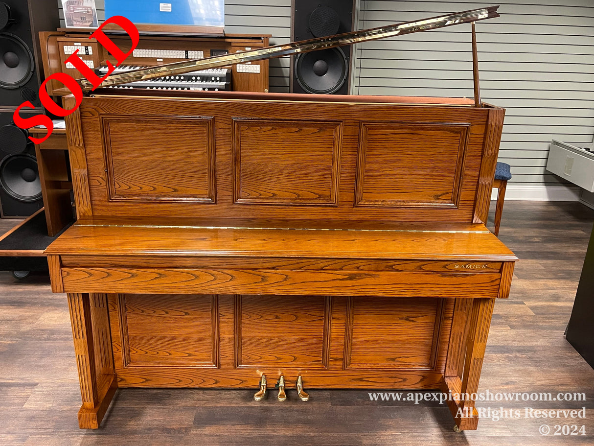 A brown Samick upright piano with a lifted top lid, exposing the strings and piano hammers, placed in a showroom with a wooden floor and speakers in the background.