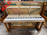 A Samick upright piano with open lid, showing internal strings and hammers, with a natural wood finish set against a showroom floor.