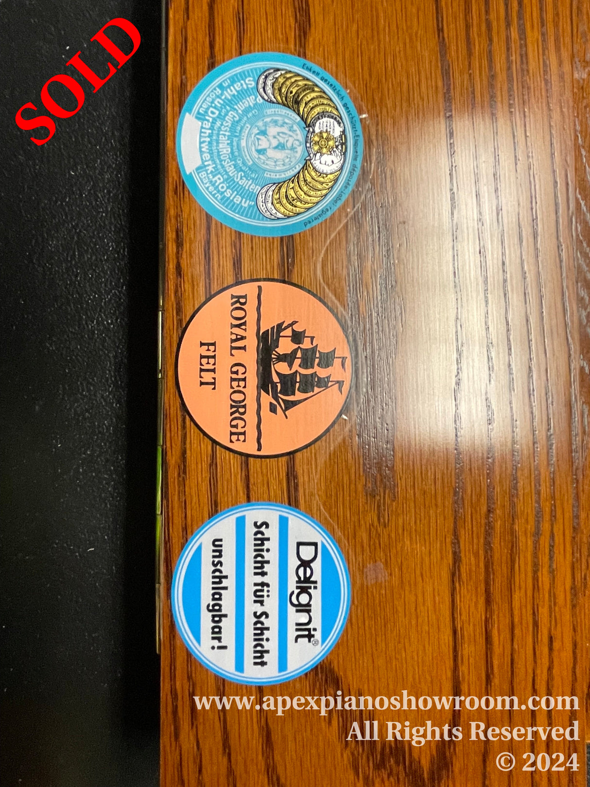 A wooden piano surface with three circular stickers — top sticker showcases piano hammers, middle sticker displays a silhouette of a grand piano, and the bottom sticker features piano tuning tools.