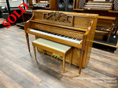 An intricately carved wooden upright piano with a matching bench, depicted in a showroom alongside other musical instruments including an organ in the background.