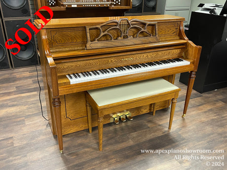 A Samick upright piano with a glossy wooden finish and decorative front panel, accompanied by a matching bench, located on a wooden floor in a music showroom setting.
