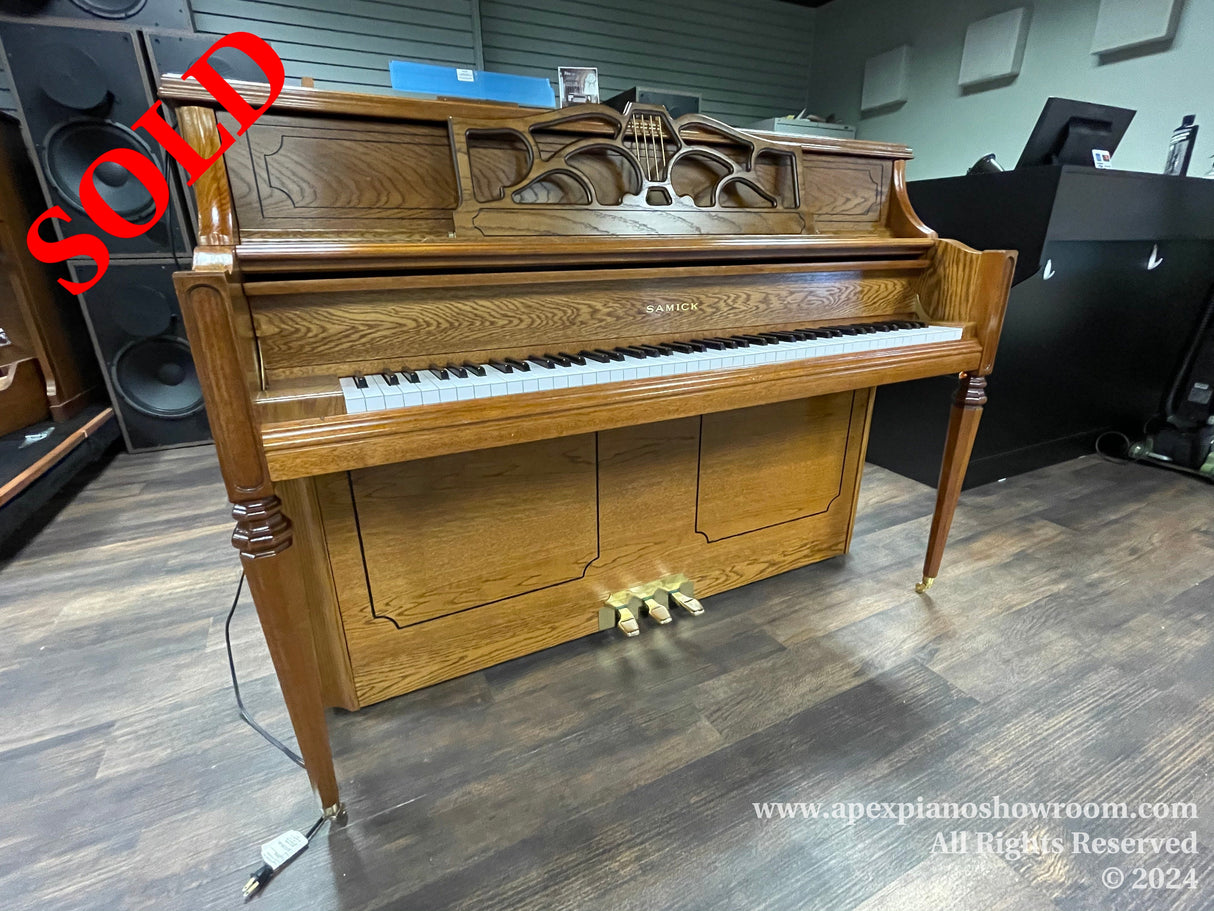 A Samick upright piano with a glossy wood finish and decorative front panel displayed in an indoor showroom, flanked by audio speakers and another piano to the right.