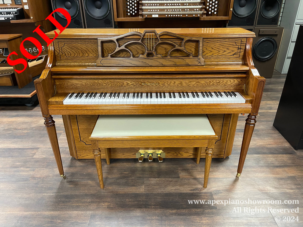 A traditional wooden upright piano with detailed carving on the front panel, ivory keys, and a matching bench set against a wooden laminate floor in a showroom.